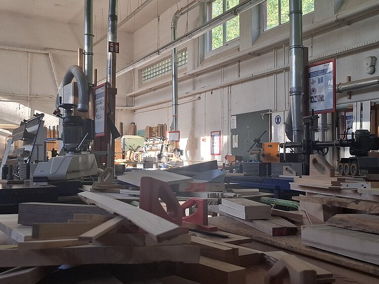 Blick durch eine Schreinerei mit Werkbänken, Holzbrettern und stationären Holzbearbeitungsmaschinen. Tageslicht fällt durch hohe Fenster und beleuchtet den Arbeitsbereich mit Werkzeugen und Materialien.