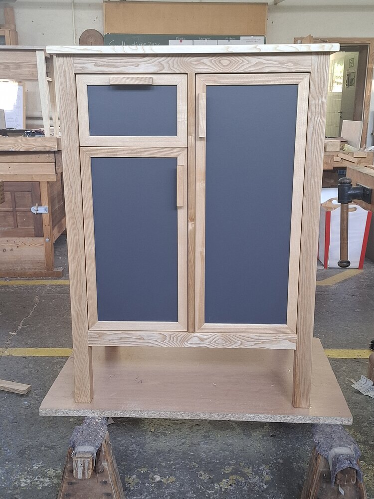 In einer Werkstatt steht ein Schrank aus hellem Holz mit drei dunkelgrauen Türen.