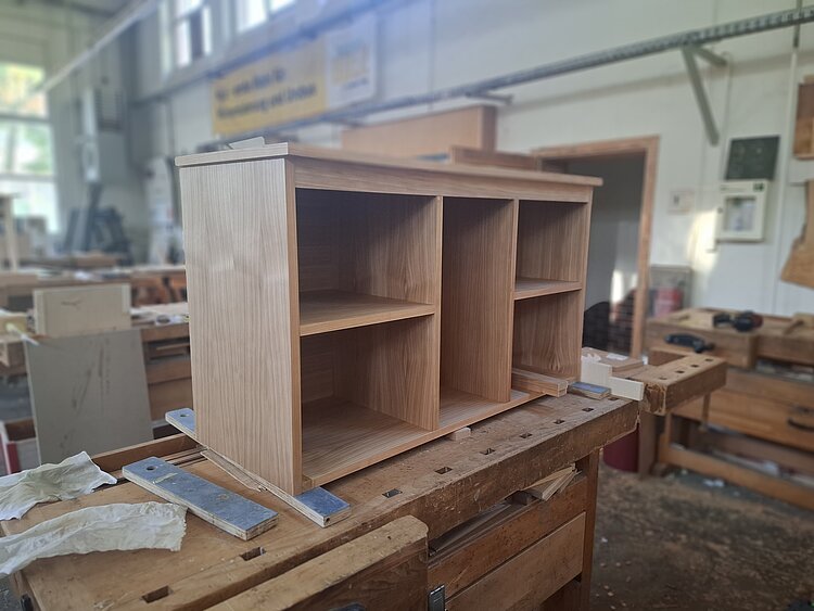 Auf einer Werkbank steht ein unfertiges Möbelstück mit offenen Fächern aus Holz.