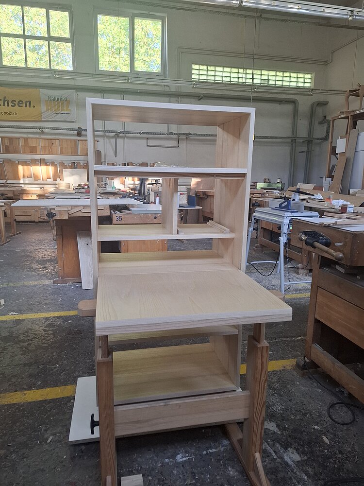 Ein unfertiges Möbelstück aus hellem Holz mit Regalfächern und ausziehbarer Arbeitsfläche steht in einer Schreinerei. Umgeben von Werkbänken und Maschinen zeigt das Bild den Entstehungsprozess eines Möbelprojekts.
