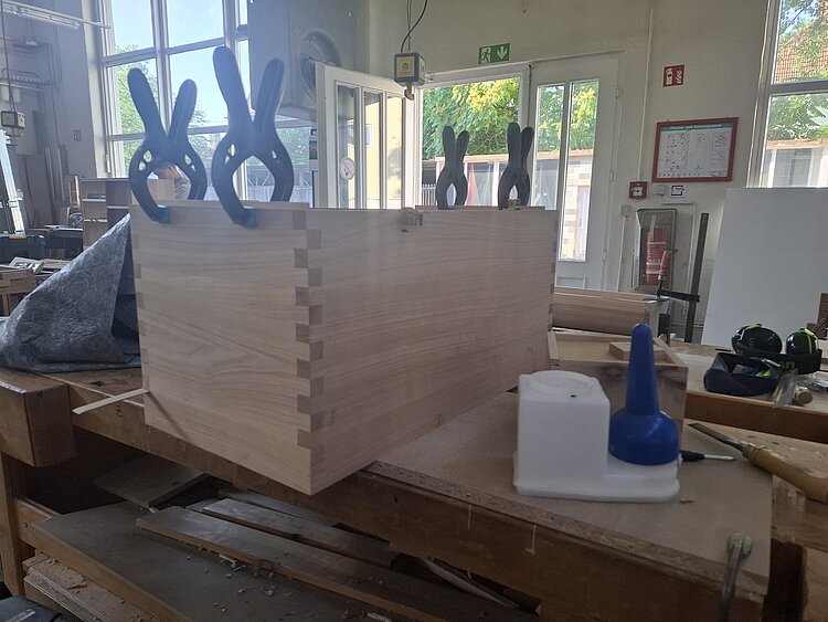 In einer Schreinerwerkstatt steht auf einem Tisch eine Holzkiste mit großen Klammern