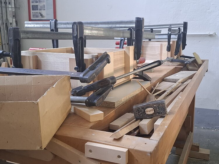 Auf einer Werkbank liegen Zwingen und Holzbauteile während des Montageprozesses.