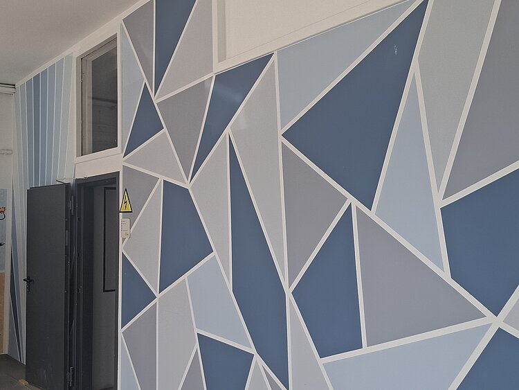 Eine Wand ist mit Dreiecken in verschiedenen Blau- und Grautönen bemalt, links sind eine offene Tür und ein Fenster zu sehen.