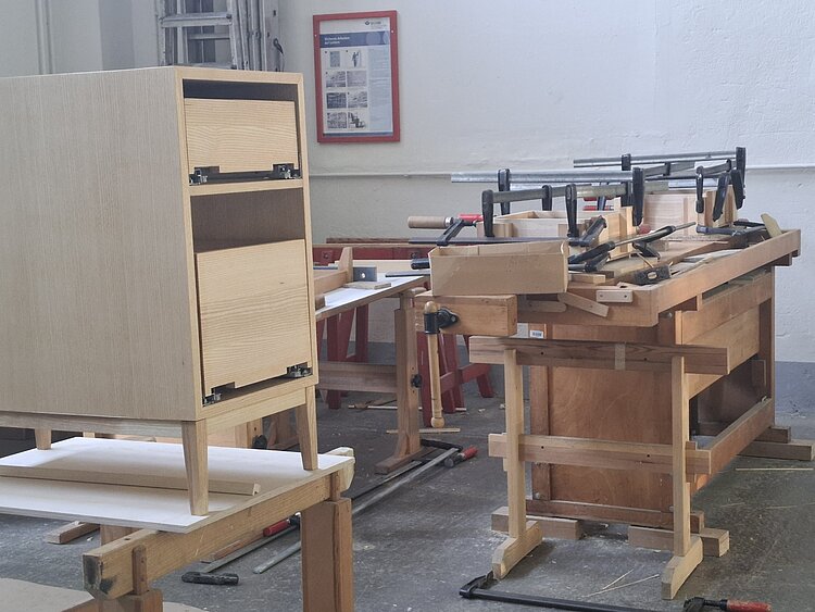 Ein teilweise fertiggestellter Holzschrank mit schmalen Schubladen steht auf einer Werkbank in einer Schreinerei. Im Hintergrund befinden sich weitere Werkbänke, Zwingen und Holzbauteile während des Montageprozesses.
