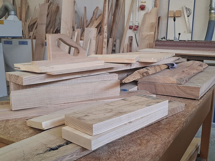 Auf einer Werkbank liegen verschiedene Holzstücke und Bretter, im Hintergrund lehnen weitere Holzbretter an der Wand